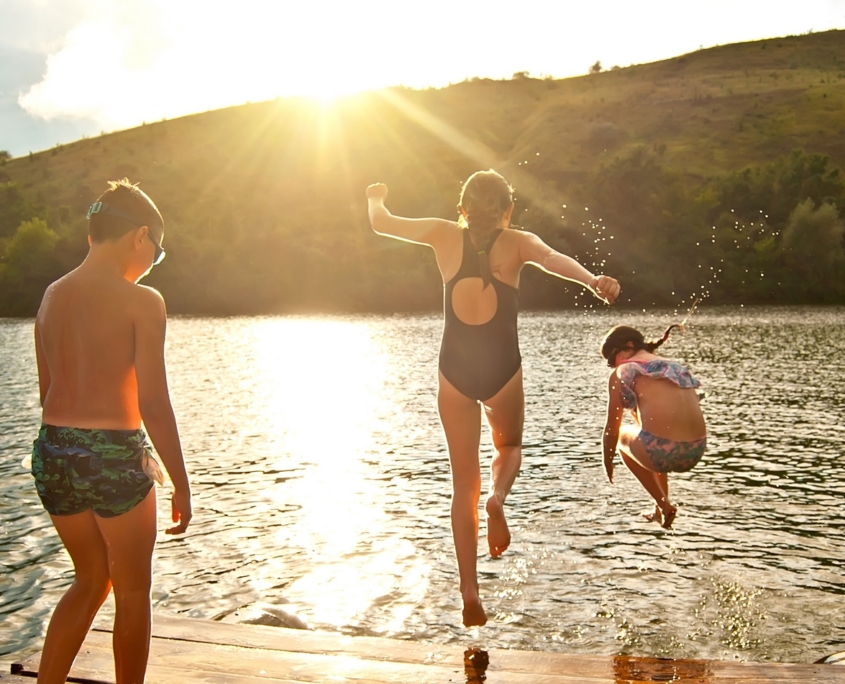 Children Jump Into The Water From The Pier. Camping By The Water